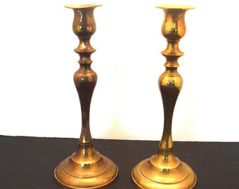 May include: Two vintage brass candlesticks. Each candlestick has a round base, a slender stem with decorative details, and a cup-shaped top to hold a candle. The candlesticks are approximately 10 inches tall.