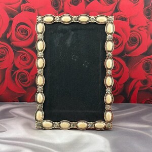 May include: A rectangular picture frame with a metallic border featuring a repeating pattern of oval cream-colored cabochons and small circular accents. The center of the frame is filled with a solid black rectangular insert.