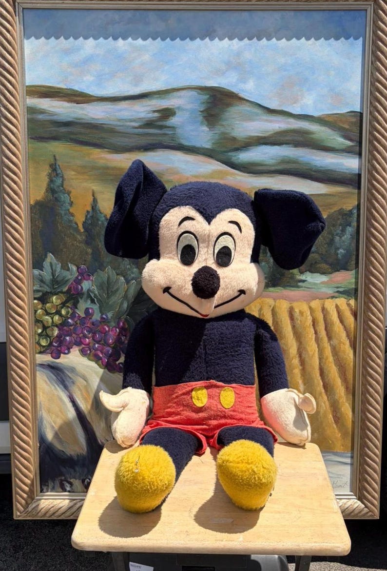 May include: A vintage Mickey Mouse plush toy with a navy blue body, red shorts, and yellow feet. The plush is seated in front of a framed landscape painting with a light wood frame. The painting depicts a vineyard.