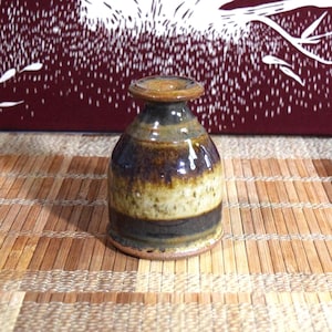 May include: A small ceramic vase with a flared rim and a rounded body. The glaze features horizontal bands of earthy tones, including dark brown, ochre, and cream, with a glossy finish that reflects light across the textured, mottled surface.