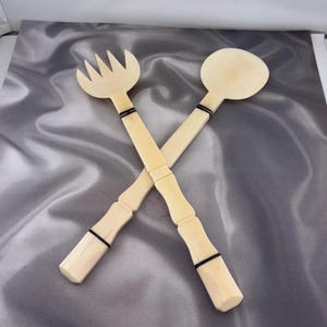 May include: A pair of light-colored wooden salad servers consisting of a fork and a spoon. Each utensil features a long handle with two distinct black bands wrapped around the shaft, and the handles have a segmented, carved design.