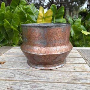 May include: A cylindrical copper pot featuring a wide, angled midsection and a flared base. The exterior surface displays a weathered, mottled patina with visible oxidation, blending warm copper tones with dark, aged metallic gray discoloration.