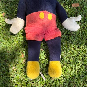 May include: A plush Mickey Mouse doll, featuring a navy blue body, red shorts with yellow button accents, white gloves, and yellow shoes. The doll is lying on a bed of green grass.