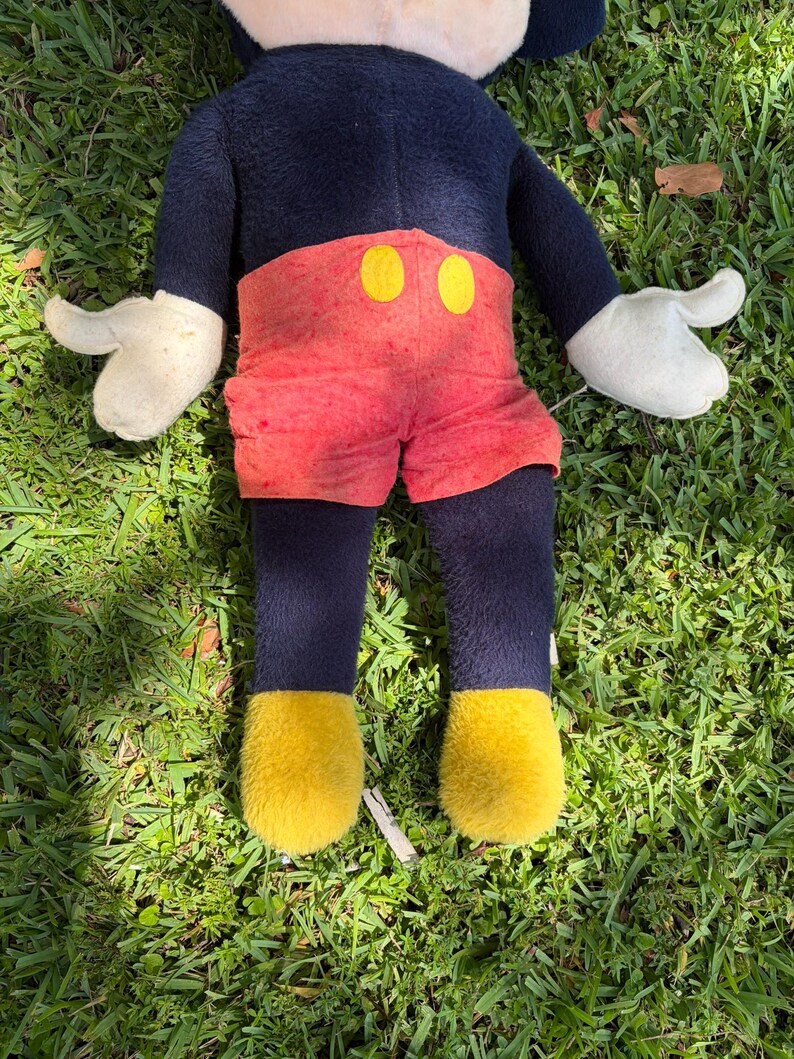 May include: A plush Mickey Mouse doll, featuring a navy blue body, red shorts with yellow button accents, white gloves, and yellow shoes. The doll is lying on a bed of green grass.