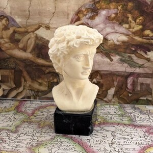 Op de afbeelding: Een kleine, gebroken witte buste van een klassieke mannelijke figuur, mogelijk David, met krullend haar en een strenge uitdrukking. De sculptuur staat op een zwarte rechthoekige basis. De achtergrond toont een muurschildering in renaissancestijl en een vintage kaart.