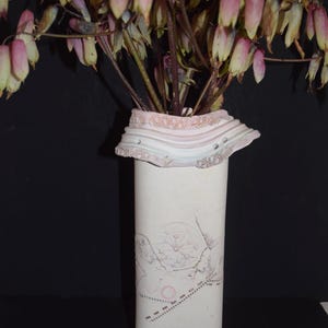 May include: A tall, rectangular ceramic vase with a matte white finish and a textured, ruffled rim in shades of pale pink and cream. The front surface features a delicate, etched geometric line drawing and a dotted pattern in faint pink ink.