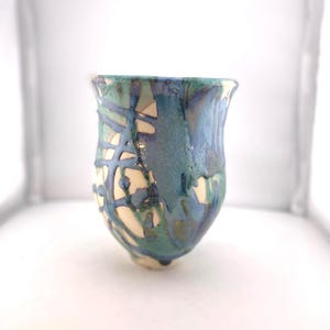 May include: A handmade ceramic cup with a unique design. The cup features a blend of blue, teal, and cream colors with a textured, dripped glaze pattern. The cup has a rounded base and a slightly flared rim.