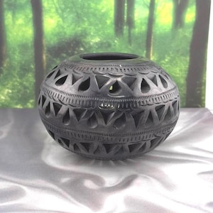May include: A spherical black ceramic vessel featuring intricate cut-out triangular patterns and horizontal bands of etched linear details. The surface has a matte finish with a wide circular opening at the top and a rounded, bulbous body shape.