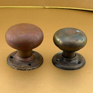May include: Two round metal door knobs with a weathered, aged patina. The left knob features a reddish-brown oxidized finish, while the right knob displays a darker, greenish-brown metallic surface. Both knobs have circular mounting plates.
