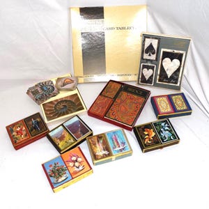 Puede incluir: Una colecci&oacute;n de juegos de cartas antiguos y un folleto de mesa de cartas de Bridge. Las cajas de cartas presentan varios dise&ntilde;os, incluyendo arreglos florales, paisajes y patrones abstractos. La paleta de colores incluye dorado, rojo, azul y verde.