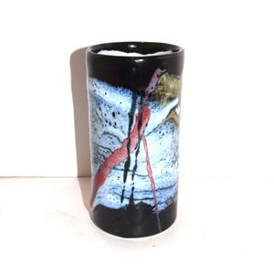 May include: A cylindrical ceramic vase with a black rim and base. The body features a white, blue, and brown speckled pattern with red and black streaks. The vase has a smooth, glossy finish and is set against a white background.
