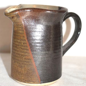 May include: A handmade ceramic pitcher with a dark brown and tan glaze. The pitcher has a curved handle and a spout for easy pouring. The design features a diagonal line of red. The pitcher is suitable for serving beverages.