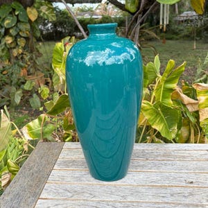 May include: A tall, smooth, teal-colored vase with a glossy finish. The vase features a rounded, bulbous body that tapers toward a narrow neck and a small, circular opening at the top. The surface is solid in color and reflects light evenly.