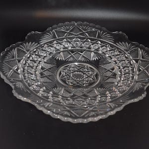 May include: A clear glass plate featuring an intricate pressed pattern of geometric stars, diamonds, and fan-shaped motifs. The circular dish has a scalloped, serrated edge and a central starburst design, showcasing a reflective, transparent finish.