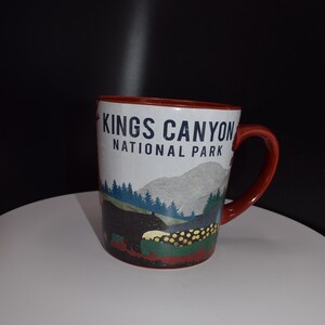May include: A red ceramic mug with a white design featuring the text "Kings Canyon National Park" and a black bear in a forest setting.