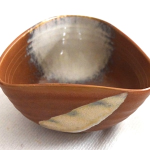 May include: A small, triangular ceramic bowl with a smooth, reddish-brown exterior and interior. The bowl features a white, glazed accent patch on the front rim and a larger, circular white glaze pattern covering the interior base and back wall.