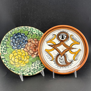 May include: Two decorative ceramic plates. One plate features a floral design with blue, yellow, and brown flowers on a green background. The other plate has a geometric design with brown and yellow accents on a white background, with an orange rim.