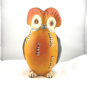 May include: A hand-painted owl figurine with a rounded body and a detailed face. The owl features orange, black, and white colors. The owl's body is primarily orange, with black accents on the wings and white details on the face.