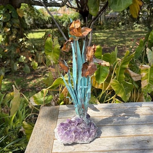 May include: A handcrafted sculpture of iris flowers. The copper-colored blooms and turquoise stems rise from a base of amethyst crystals. The sculpture is displayed on a wooden table outdoors.
