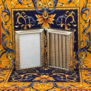 May include: A hinged, silver-colored picture frame with two rectangular openings. One side is empty, while the other contains a series of vertical, brown, wooden rods. The frame is set against a patterned background with blue, orange, and yellow floral designs.