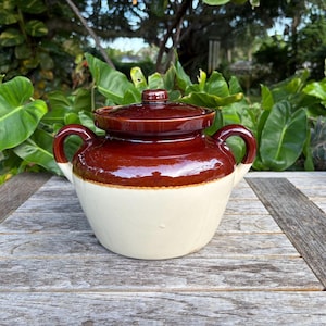 May include: A two-handled ceramic bean pot featuring a glossy dark brown glazed lid and upper body, transitioning to a smooth cream-colored base. The vessel includes two curved side handles and a matching brown lid with a central knob handle.