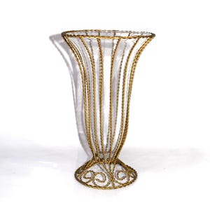 May include: A clear glass vase with a gold-colored metal wire frame. The vase has a flared top and a decorative base. The gold wire is woven in a vertical pattern, creating a cage-like design. The vase is approximately 10 inches tall.