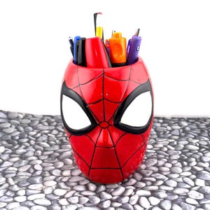 May include: A red pen holder shaped like Spider-Man's mask, with black detailing and large white eye shapes. The pen holder is filled with various pens and markers. The product is a fun and functional desk accessory.