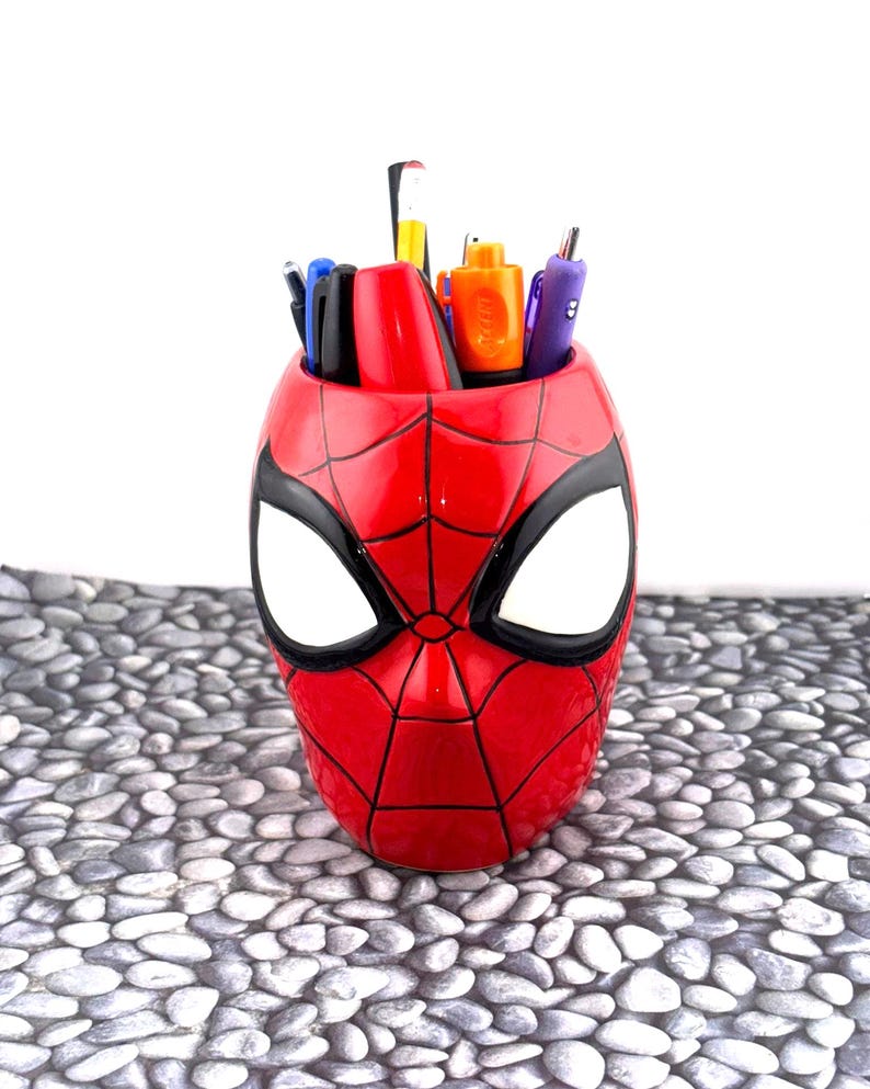 May include: A red pen holder shaped like Spider-Man's mask, with black detailing and large white eye shapes. The pen holder is filled with various pens and markers. The product is a fun and functional desk accessory.