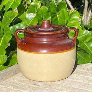 May include: A two-toned ceramic crock with a lid and two handles. The top portion of the crock and lid are a dark brown, while the bottom is a cream color. The crock is sitting on a wooden surface.
