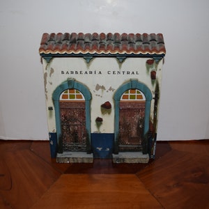 May include: A small, white building with a red tile roof and blue trim. The building has two doors with arched windows and the words "BARBEARIA CENTRAL" written above the doors.