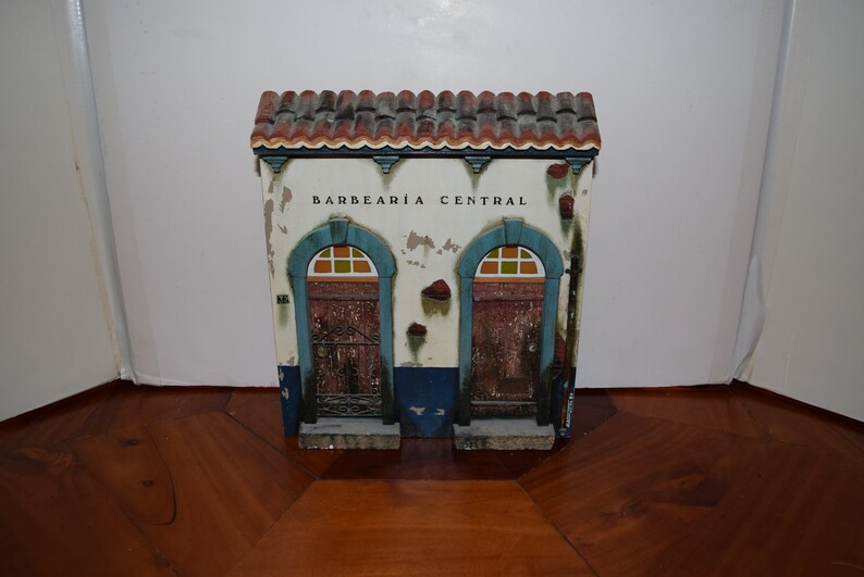 May include: A small, white building with a red tile roof and blue trim. The building has two doors with arched windows and the words "BARBEARIA CENTRAL" written above the doors.