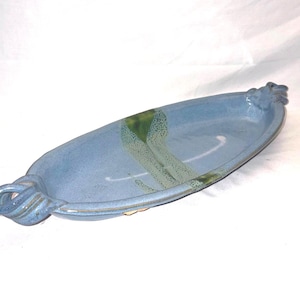 May include: A handmade, oval-shaped ceramic serving dish in a light blue glaze. The dish features a decorative handle on each end and green and white accents. The overall design is simple and elegant, suitable for serving appetizers or small dishes.
