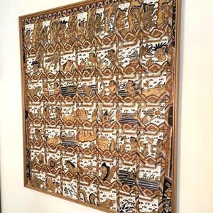 Op de afbeelding: Een ingelijst kunstwerk met een herhalend patroon van figuren en dieren in bruin, zwart en wit. Het kunstwerk is ingesloten in een houten lijst, wat een vleugje warmte en elegantie toevoegt. Het thema is een cultureel of historisch verhaal.