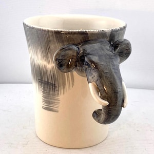 May include: A ceramic mug with an elephant trunk handle. The mug is off-white with a gray and black painted design on the upper portion. The elephant trunk handle is gray with white tusks.