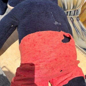 May include: A well-worn plush toy featuring a dark blue body and red shorts. The toy shows signs of wear, including a hole in the shorts. The toy is positioned on a light-colored wooden surface.