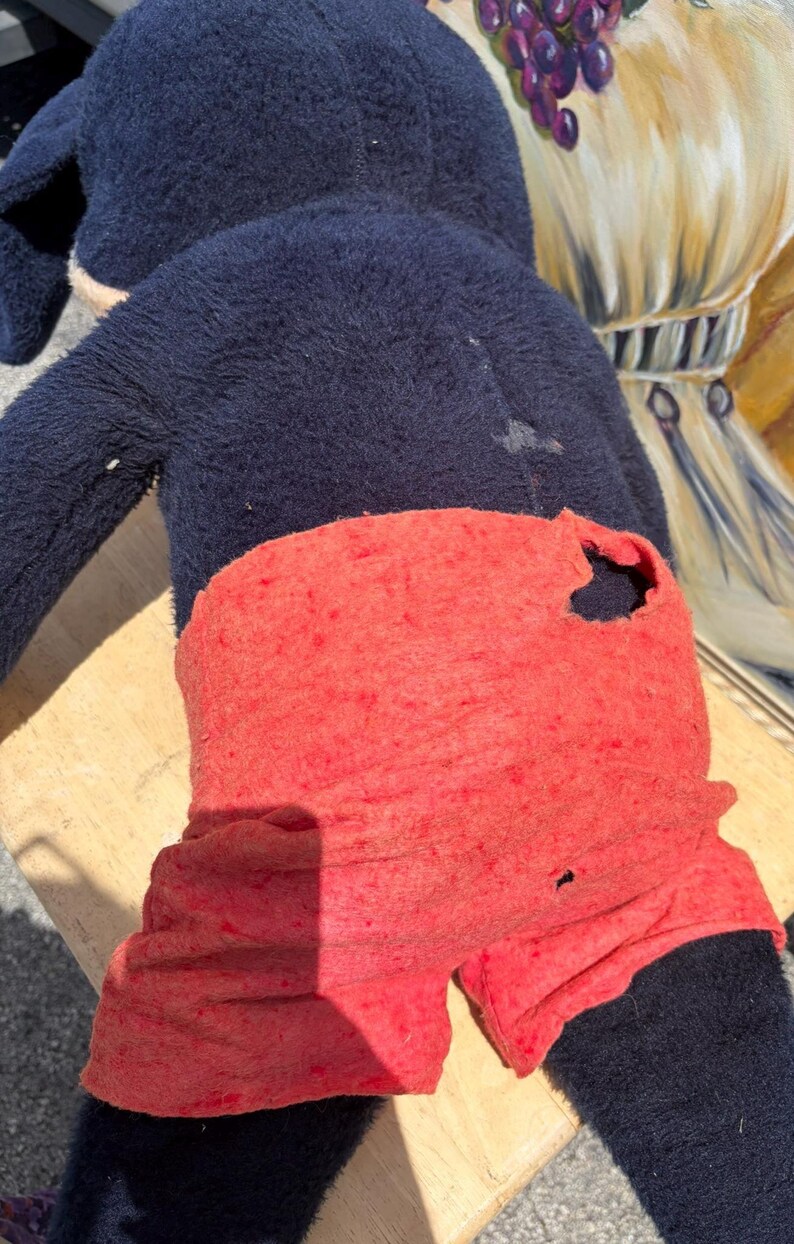 May include: A well-worn plush toy featuring a dark blue body and red shorts. The toy shows signs of wear, including a hole in the shorts. The toy is positioned on a light-colored wooden surface.