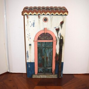 May include: A decorative wall plaque shaped like a building facade with a terracotta-colored tiled roof, a weathered white wall with blue trim, a pink arched door frame, and a dark teal wooden door featuring the number 46 and metal accents.