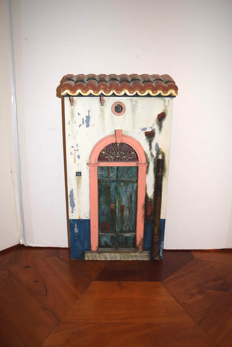 May include: A decorative wall plaque shaped like a building facade with a terracotta-colored tiled roof, a weathered white wall with blue trim, a pink arched door frame, and a dark teal wooden door featuring the number 46 and metal accents.