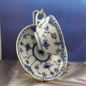 May include: A porcelain teacup and saucer set with a blue floral pattern. The cup is suspended on a gold-colored metal stand. The design features intricate blue floral motifs on a white background, creating an elegant aesthetic.