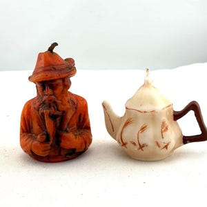 May include: Two decorative candles: one shaped like an old man with a long beard and hat, and the other a small teapot. The man candle is orange-brown, and the teapot is off-white with brown accents.