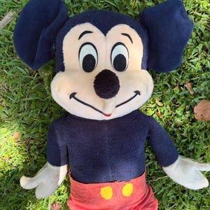 May include: A vintage Mickey Mouse plush toy. The plush has a navy blue body, large black ears, and a cream-colored face. It wears red shorts with yellow buttons. The toy is lying on green grass.