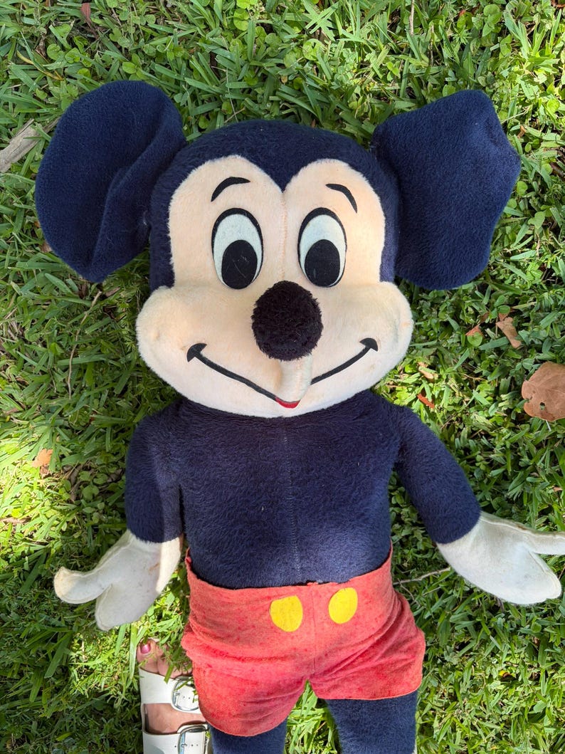 May include: A vintage Mickey Mouse plush toy. The plush has a navy blue body, large black ears, and a cream-colored face. It wears red shorts with yellow buttons. The toy is lying on green grass.