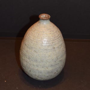May include: A small, light gray ceramic vase with a textured surface. The vase has a rounded, egg-shaped body and a narrow opening at the top.