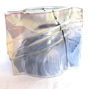 May include: A handcrafted ceramic vase with a unique, asymmetrical shape. The vase features a blend of colors, including blue, gray, and yellow, with a glossy finish. The design includes horizontal ridges and a dark, vertical accent.