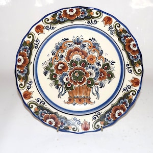 K&ouml;nnte beinhalten: Ein dekorativer Keramikteller mit gewelltem Rand. Der Teller zeigt ein zentrales Blumenkorb-Design in Braun-, Orange-, Blau- und Gr&uuml;nt&ouml;nen, umgeben von einem blauen Rand und floralen Akzenten. Der Teller hat einen Durchmesser von etwa 25 cm.