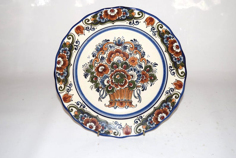 K&ouml;nnte beinhalten: Ein dekorativer Keramikteller mit gewelltem Rand. Der Teller zeigt ein zentrales Blumenkorb-Design in Braun-, Orange-, Blau- und Gr&uuml;nt&ouml;nen, umgeben von einem blauen Rand und floralen Akzenten. Der Teller hat einen Durchmesser von etwa 25 cm.