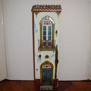 May include: A white, two-story, miniature building with a red tile roof, a blue door, and a balcony with a wrought iron railing. The building has a distressed paint finish with blue and white accents.