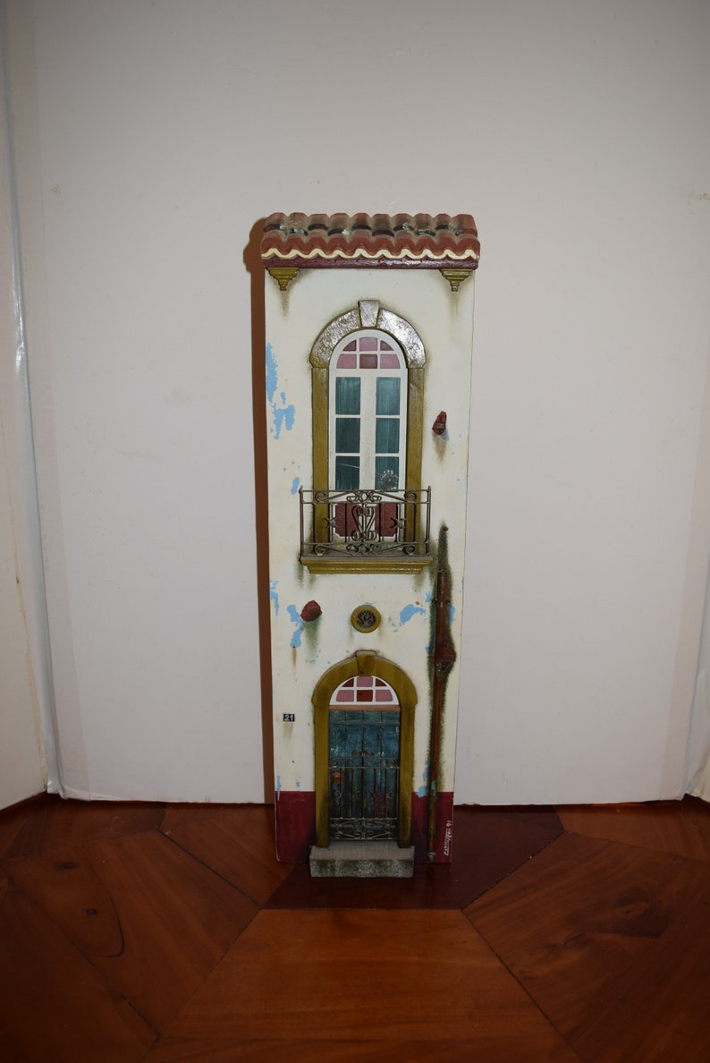 May include: A white, two-story, miniature building with a red tile roof, a blue door, and a balcony with a wrought iron railing. The building has a distressed paint finish with blue and white accents.