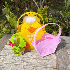 May include: Three Easter-themed felt baskets in the image. A yellow chick basket, a pink bunny basket, and a green basket with a butterfly and floral design. The baskets have handles and are set on a wooden surface.