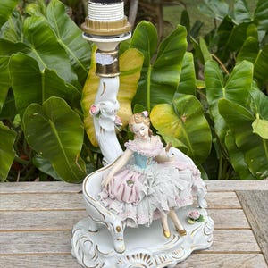 May include: A white porcelain lamp base featuring a seated figurine in a ruffled dress. The figurine is in shades of pink, blue, and white, with gold accents. The lamp base has a decorative design and a brass lamp holder.